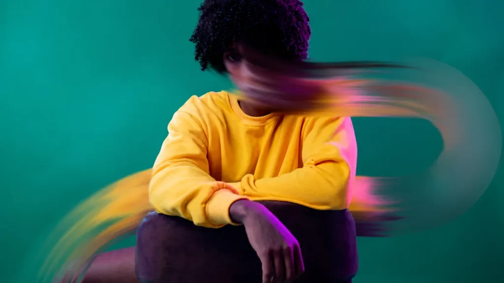 A person in a yellow sweatshirt sits against a teal background while their head blurs in motion, suggesting anxious, racing thoughts.