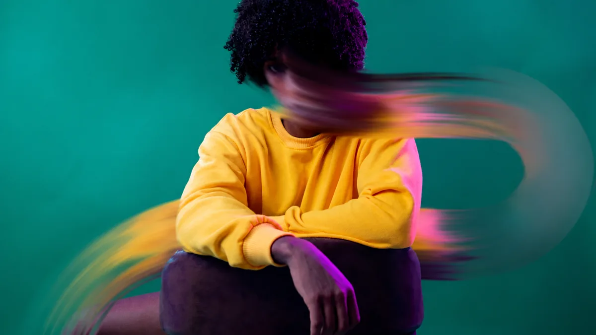 Therapist Aid Worksheets For Anxiety: How to Choose and Use Them 2 A person in a yellow sweatshirt sits against a teal background while their head blurs in motion, suggesting anxious, racing thoughts.
