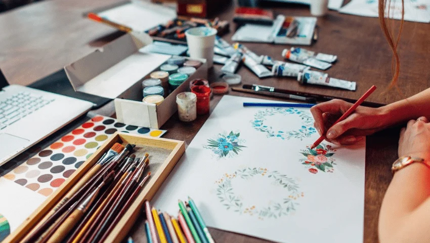 Person painting a floral watercolor beside brushes and a paint palette.
